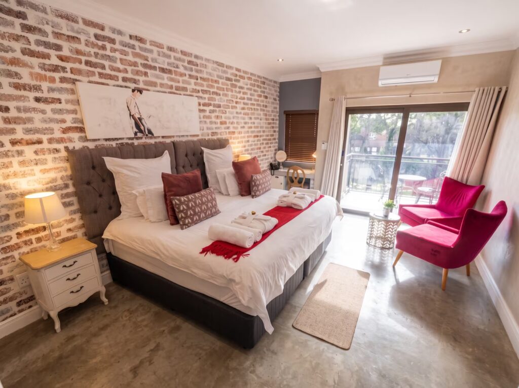 A luxurious, sunlit boutique hotel room featuring a plush king-size bed with crisp white linens and red accent pillows against an exposed brick wall. In the foreground, two striking magenta velvet armchairs sit near sliding glass doors that open to a balcony. The room is styled with polished concrete floors, a large framed artwork over the bed, vintage-style white nightstands, and warm ambient lighting.