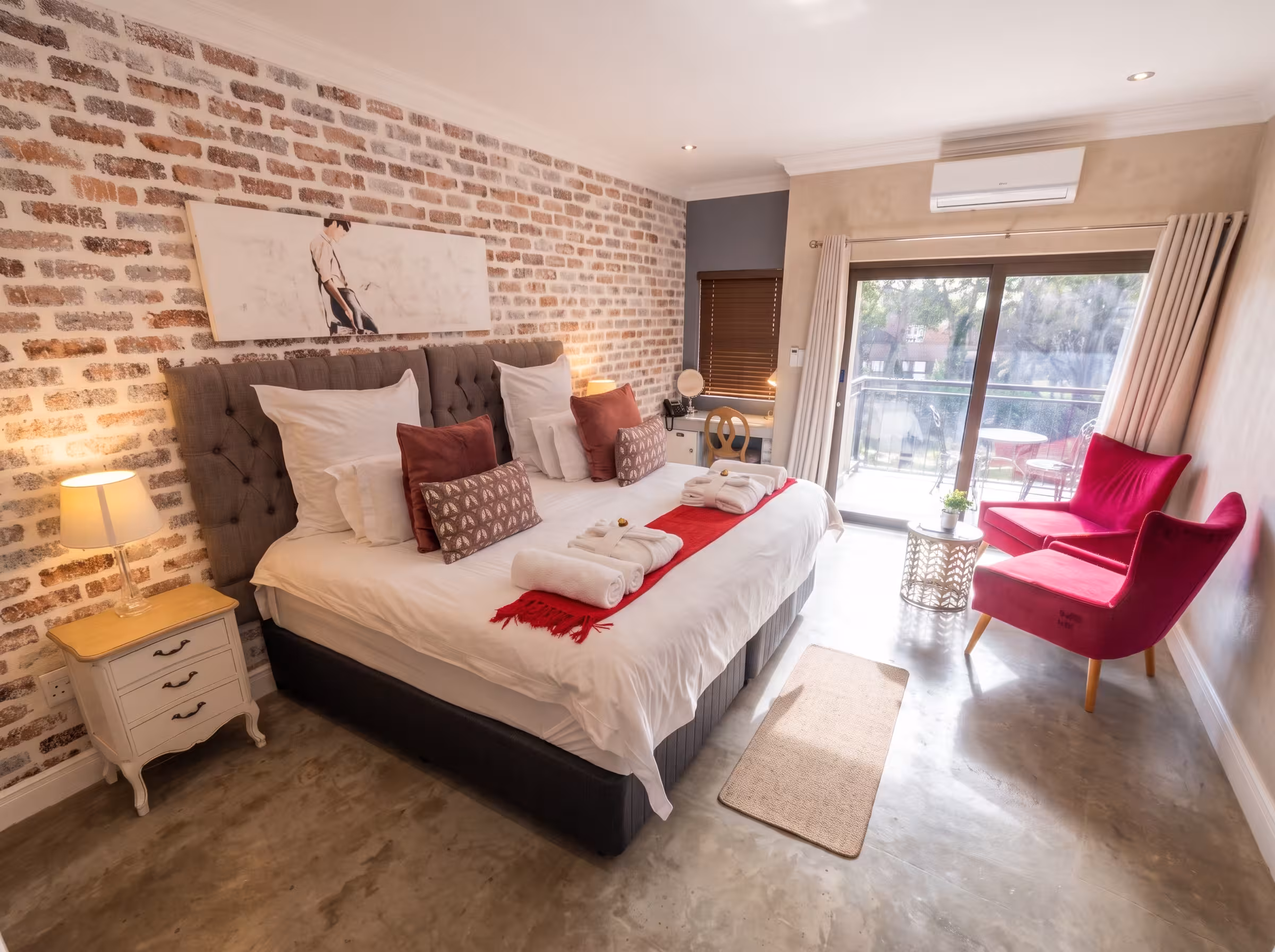 A luxurious, sunlit boutique hotel room featuring a plush king-size bed with crisp white linens and red accent pillows against an exposed brick wall. In the foreground, two striking magenta velvet armchairs sit near sliding glass doors that open to a balcony. The room is styled with polished concrete floors, a large framed artwork over the bed, vintage-style white nightstands, and warm ambient lighting.